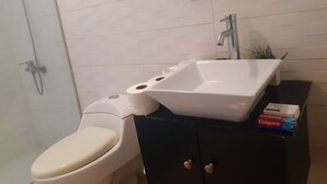Une salle de bain avec des toilettes blanches, un lavabo avec un miroir au-dessus et une douche. On peut voir une boîte de dentifrice Colgate sur le comptoir.