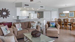 A spacious living room with a ceiling fan, a glass coffee table, and a dining area with wooden chairs and a white table. - Sandpiper Condominiums 902, Panoramic Gulf Views w/ direct access to Mustang Island/Port A beaches. (Port Aransas)