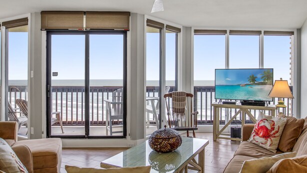 A spacious living room with a large television, comfortable seating, and a view of the ocean through large windows. - Sandpiper Condominiums 902, Panoramic Gulf Views w/ direct access to Mustang Island/Port A beaches. (Port Aransas)