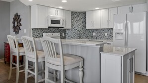 A kitchen with white cabinets, a microwave, a refrigerator, and a bar area with three chairs. - Sandpiper Condominiums 902, Panoramic Gulf Views w/ direct access to Mustang Island/Port A beaches. (Port Aransas)