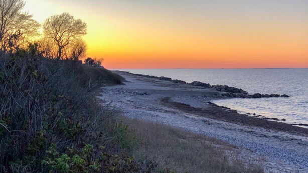 Eine Küstenlandschaft mit felsiger Uferlinie, kargen Pflanzen und einem Sonnenuntergang über dem Wasser.
