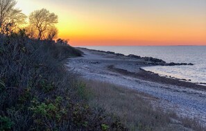 Rannikkomaisema, jossa on kivinen rantaviiva, niukasti kasvillisuutta ja auringonlasku veden yllä.