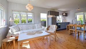 Ein gemütlicher, hell eingerichteter Raum mit Holzboden, einem weißen Sofa, Korbsesseln, einem Esstisch und einer Küchenzeile mit Schränken und einer Spüle.