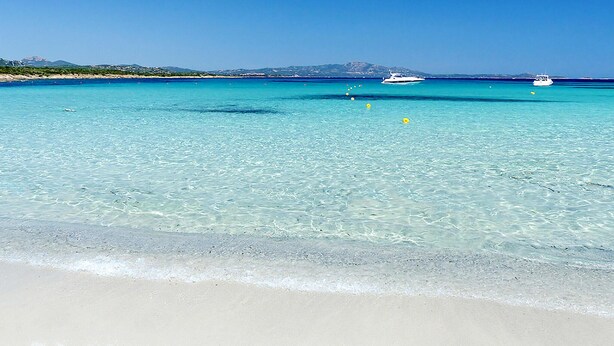 Une plage avec une eau turquoise et limpide, un rivage sablonneux et des bateaux à l’horizon.