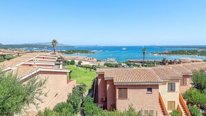 Une zone résidentielle côtière avec des maisons aux toits de tuiles rouges, une mer d’un bleu limpide et des bateaux visibles au loin.