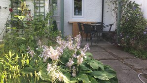 Ein Garten mit einem Holztisch und Stühlen, ein weißes Gebäude mit grünen Akzenten sowie verschiedene Pflanzen und Blumen.