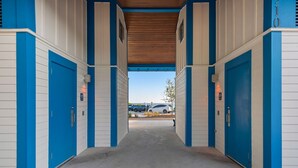 Un pasillo cubierto que conduce a un edificio con revestimiento azul y blanco, suelo de concreto y una vista despejada de automóviles estacionados al fondo.