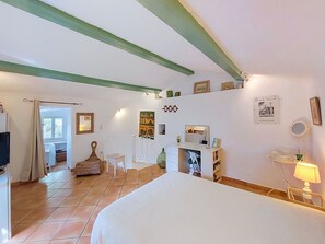Een slaapkamer met een bed, een bureau, een stoel en een klein tafeltje. Er is een raam met gordijnen, een muur met ingelijste foto’s en een betegelde vloer.