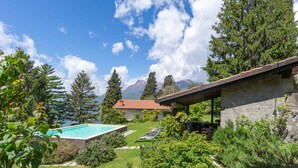Une piscine avec vue sur les montagnes, un bâtiment en pierre doté d’un toit et un jardin où poussent différentes espèces de plantes.