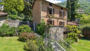 Un bâtiment à deux étages, avec une base en pierre et une partie supérieure de couleur beige, entouré d’une végétation luxuriante et d’un mur en pierre.