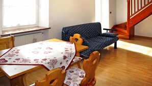 Een gezellige kamer met een houten tafel, een blauw gestreept bankje en een houten stoel. Er is een raam met gordijnen, een houten trap en een hangende lamp.