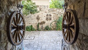 A stone courtyard with two large wooden wheels, a cobblestone path, and a stone wall with a decorative metal gate.