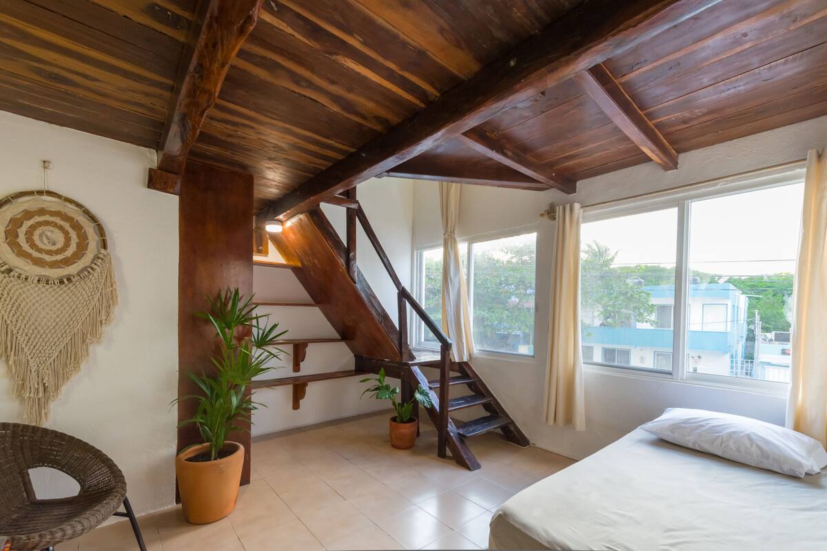 A room with a wooden ceiling, a bed, a staircase, and a view through a window.