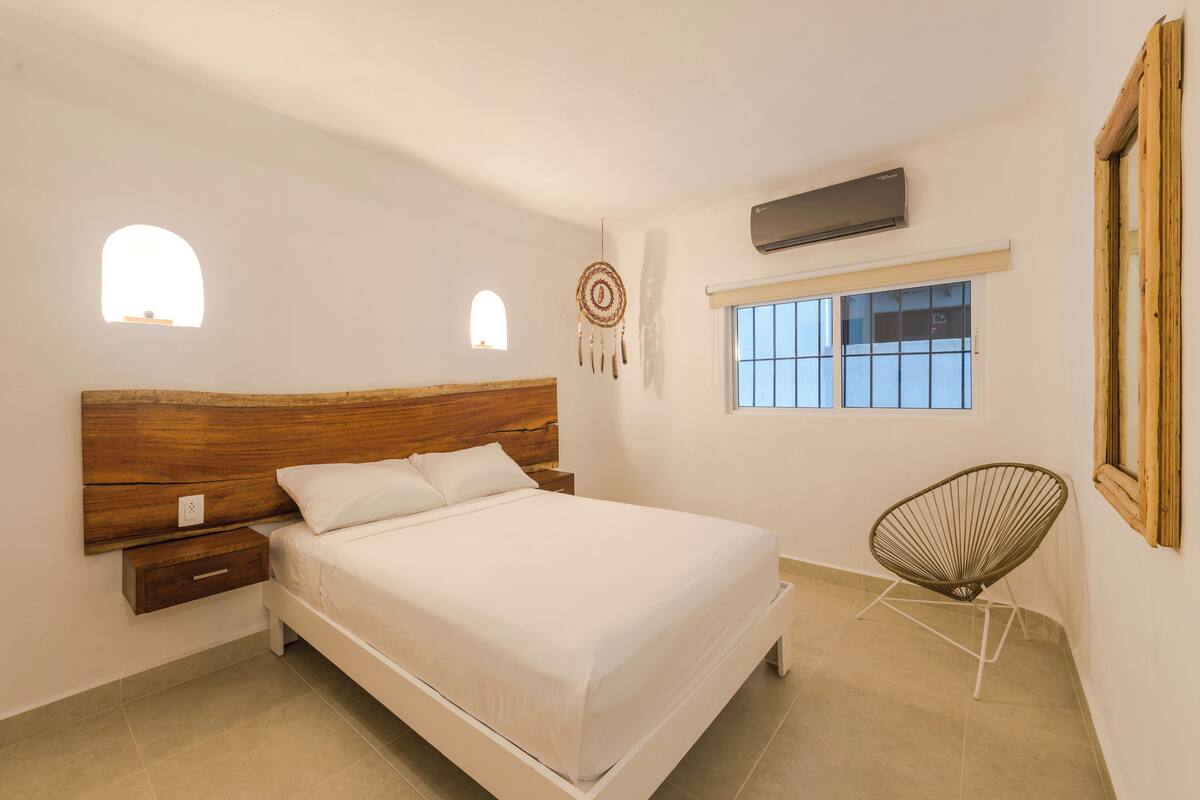 A bedroom with a wooden headboard, a single bed, a nightstand, a dreamcatcher, a window, and a chair.