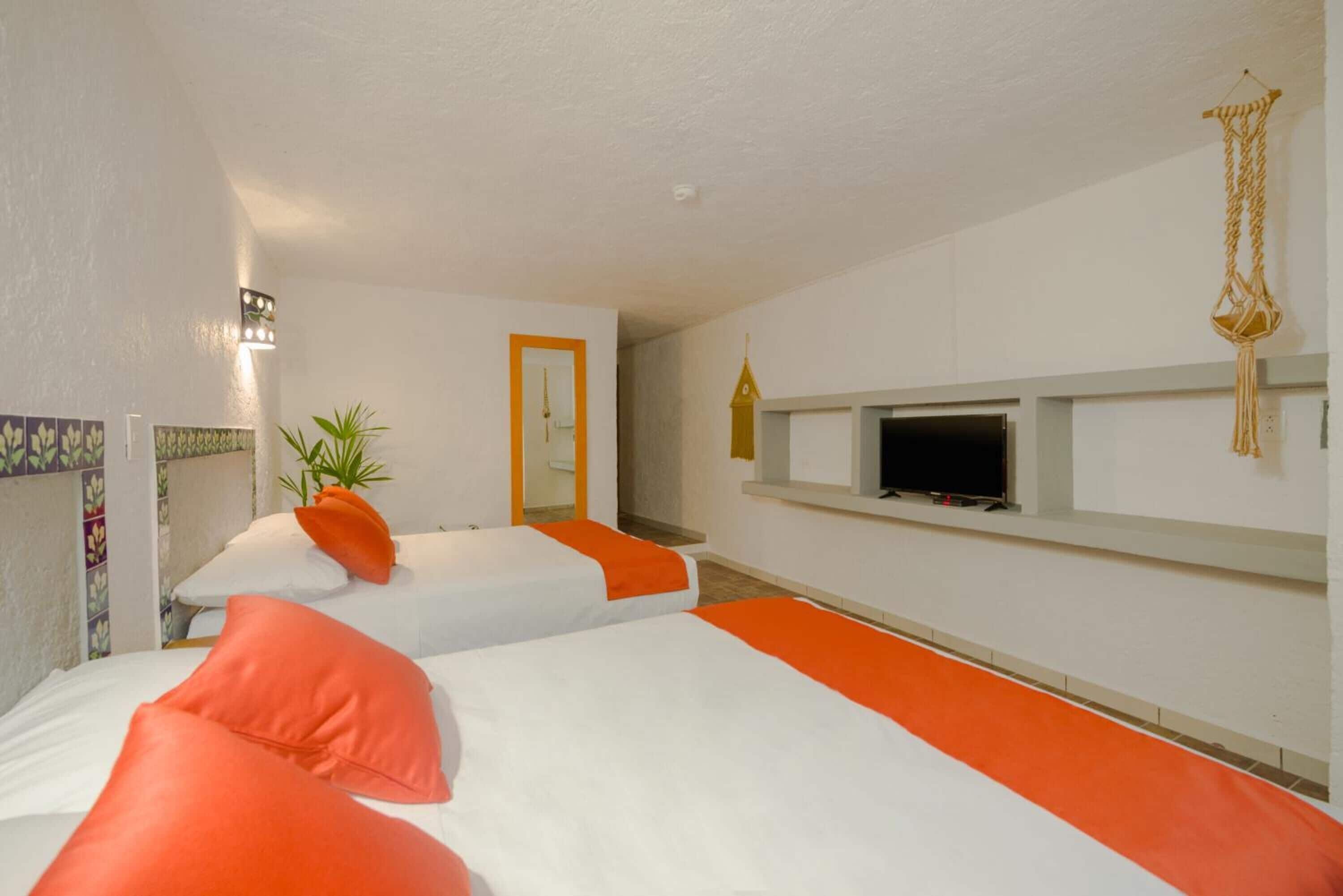 Ein Hotelzimmer mit zwei Betten, jeweils mit weißer und orangefarbener Bettwäsche, einem Fernseher auf einem Regal, einer Pflanze und einer Makramee-Girlande an der Decke.