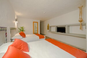 Habitación de hotel con dos camas, cada una con ropa de cama blanca y naranja, un televisor en un estante, una planta y un macramé colgando del techo.