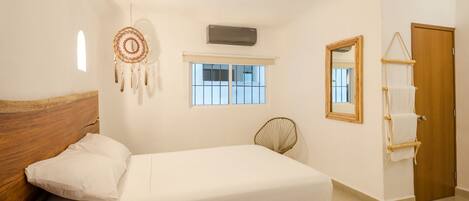 Une chambre avec une tête de lit en bois, un lit simple, un attrape-rêves, une fenêtre, un climatiseur, un miroir et une porte en bois.
