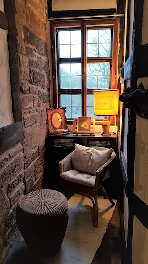 A cozy room with a stone wall, a wooden chair, a wicker ottoman, and a desk with a lamp and framed picture.