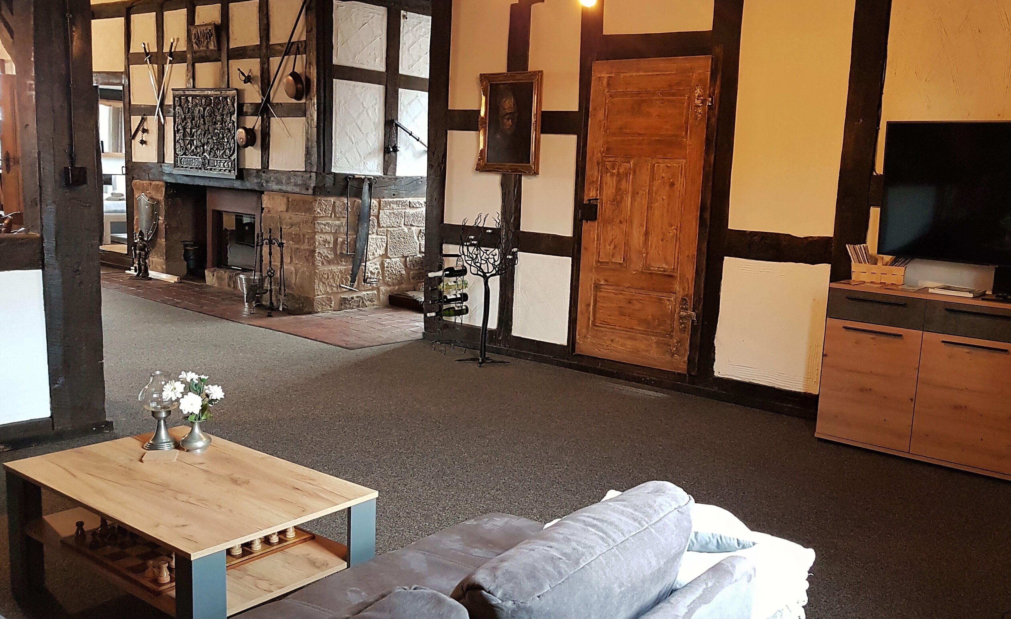 A traditional room with exposed wooden beams, a stone fireplace, a wooden table with chess pieces, and a grey sofa.