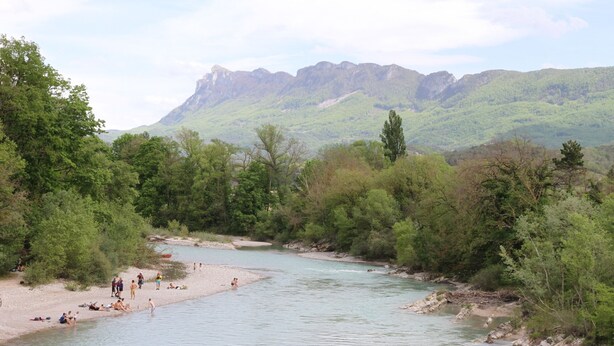 Een rivier met mensen die ontspannen op de oevers, omringd door weelderige begroeiing en bergen op de achtergrond.