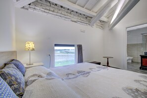 A bedroom with a large bed, a bedside lamp, a view of the sea through a window, and a small table.