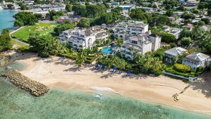 An aerial view of a coastal resort with a large building, a swimming pool, and a beach area with umbrellas. - Beachfront Retreat with Pool: Schooner Bay 112 (Speightstown)
