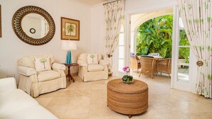 A spacious living room with beige sofas, a round wicker ottoman, and a large mirror. - Beachfront Retreat with Pool: Schooner Bay 112 (Speightstown)