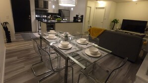 Un espace repas moderne avec une table en verre pour quatre personnes, entourée de chaises transparentes. On aperçoit, au fond, une cuisine équipée d’appareils électroménagers en acier inoxydable.