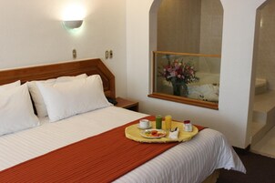 Habitación de hotel con cama, una bandeja de desayuno, un cuadro y baño visible a través de un arco.