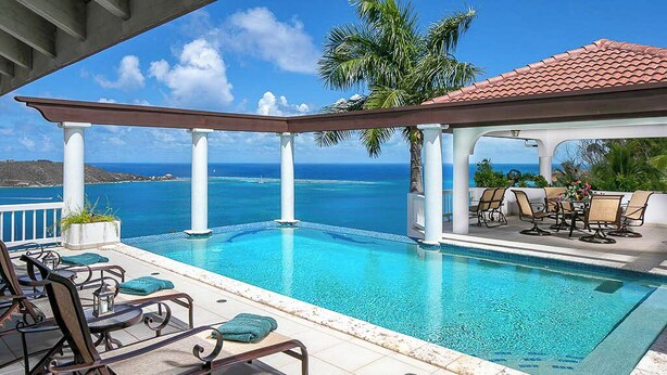 Un espace aménagé au bord de la piscine, avec des chaises longues et un coin salon couvert offrant une vue sur un océan d’un bleu limpide.