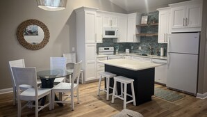 A modern kitchen with white cabinetry, a central island, and a dining area with white chairs. - Once Upon a Tide: FULL RENO for 2026! Oceanfront, Pool, Linens (Indian Beach)