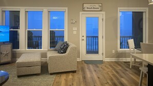 A living room with a television, a sofa, a dining table, and a door leading to a balcony. - Once Upon a Tide: FULL RENO for 2026! Oceanfront, Pool, Linens (Indian Beach)