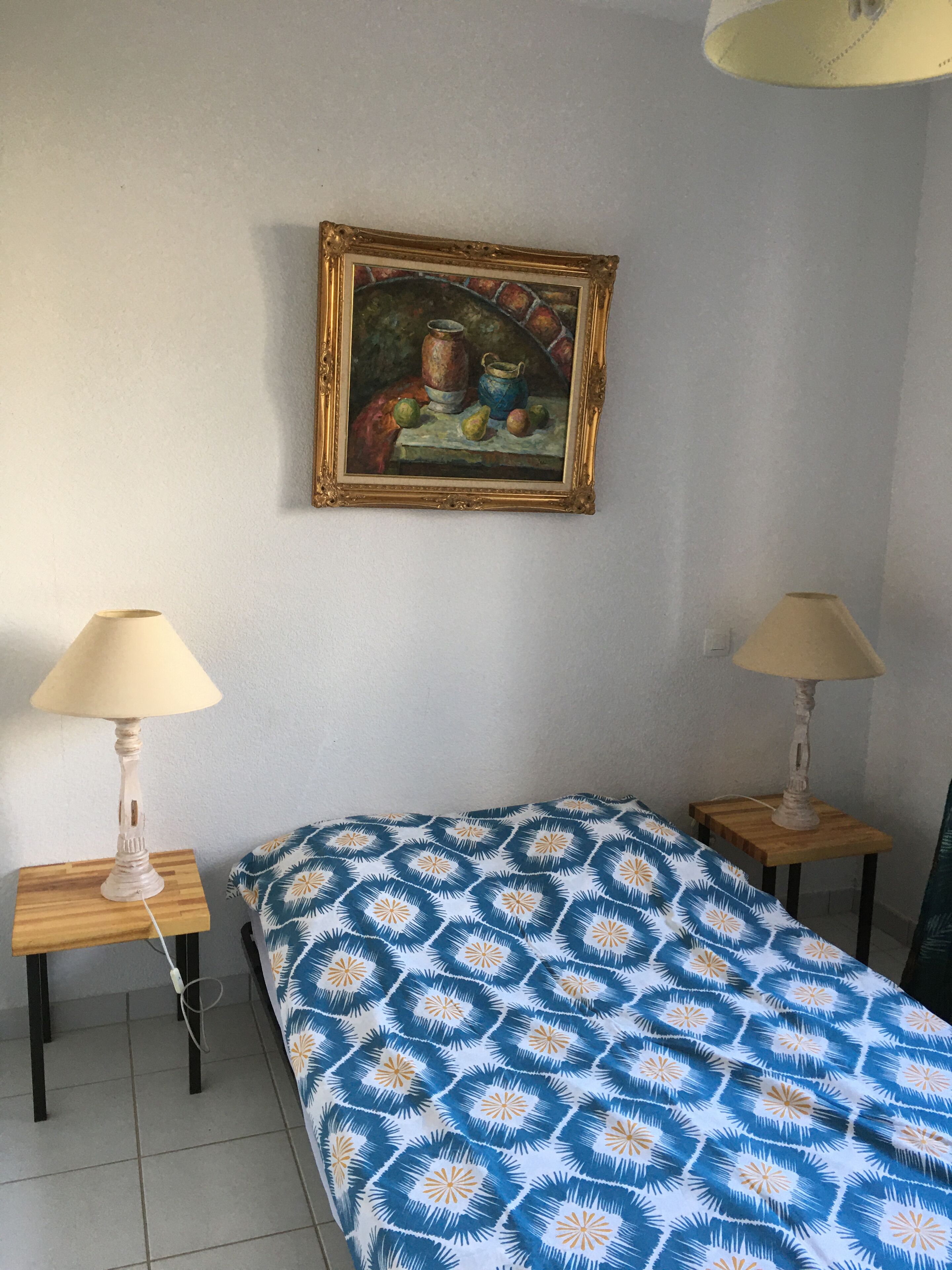 Un dormitorio con una cama, dos mesitas de noche, una lámpara sobre cada una, un cuadro en la pared y una colcha estampada.