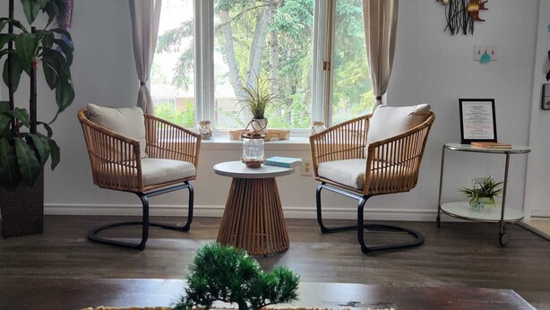 Une pièce comprenant deux fauteuils en osier avec des coussins blancs, une petite table ronde surmontée d’un vase en verre et une plante en pot.