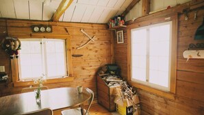 A rustic cabin interior with wooden walls, a round dining table, two windows, and a small kitchenette area.
