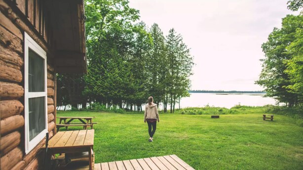 A wooden cabin with a deck and picnic table, surrounded by a grassy area and trees, overlooking a body of water.