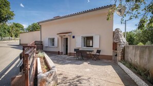 Un edificio de una sola planta con fachada color beige y techo de tejas. Cuenta con un pequeño patio con mesa y sillas, y un muro de piedra a la derecha.