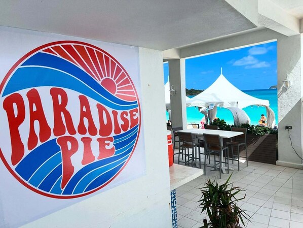 A sign with 'PARADISE PIE' and a view of a beach with umbrellas and a tent.
