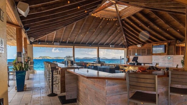 A wooden bar area with a view of the sea, a TV, and a seating area with chairs. - Lovely Sapphire Beachfront Condo - Snorkel Crystal Cove - Paradise ! (St Thomas)
