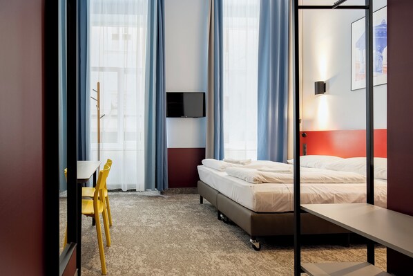 A hotel room with a bed, a desk with a chair, a TV mounted on the wall, and a window with curtains.