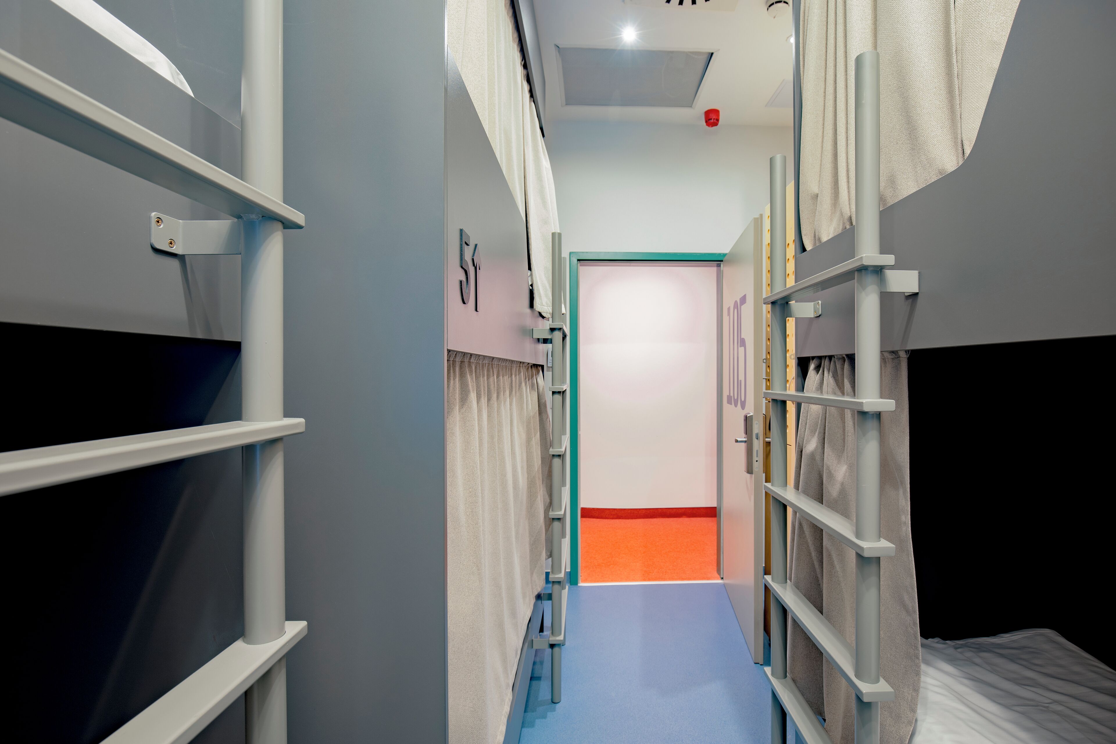 A narrow corridor with bunk beds on either side, a door at the end, and a blue carpet.