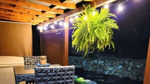 An outdoor wooden deck with a table and chairs, a hanging fern, and a stone wall with a small water feature.