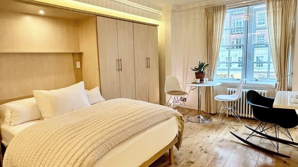Une chambre d’hôtel avec un grand lit, un bureau avec une chaise, une petite table avec une plante et une fenêtre avec des rideaux.
