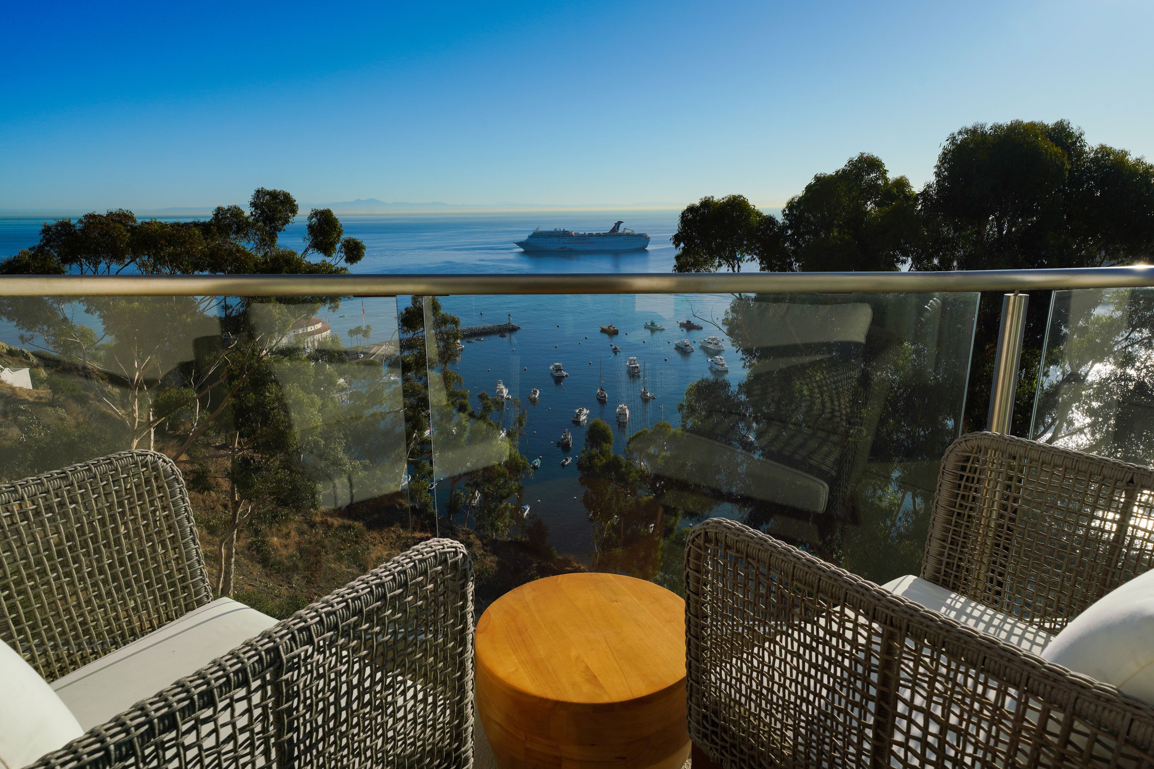 A balcony with wicker furniture and a wooden table, overlooking a marina with boats and a cruise ship.