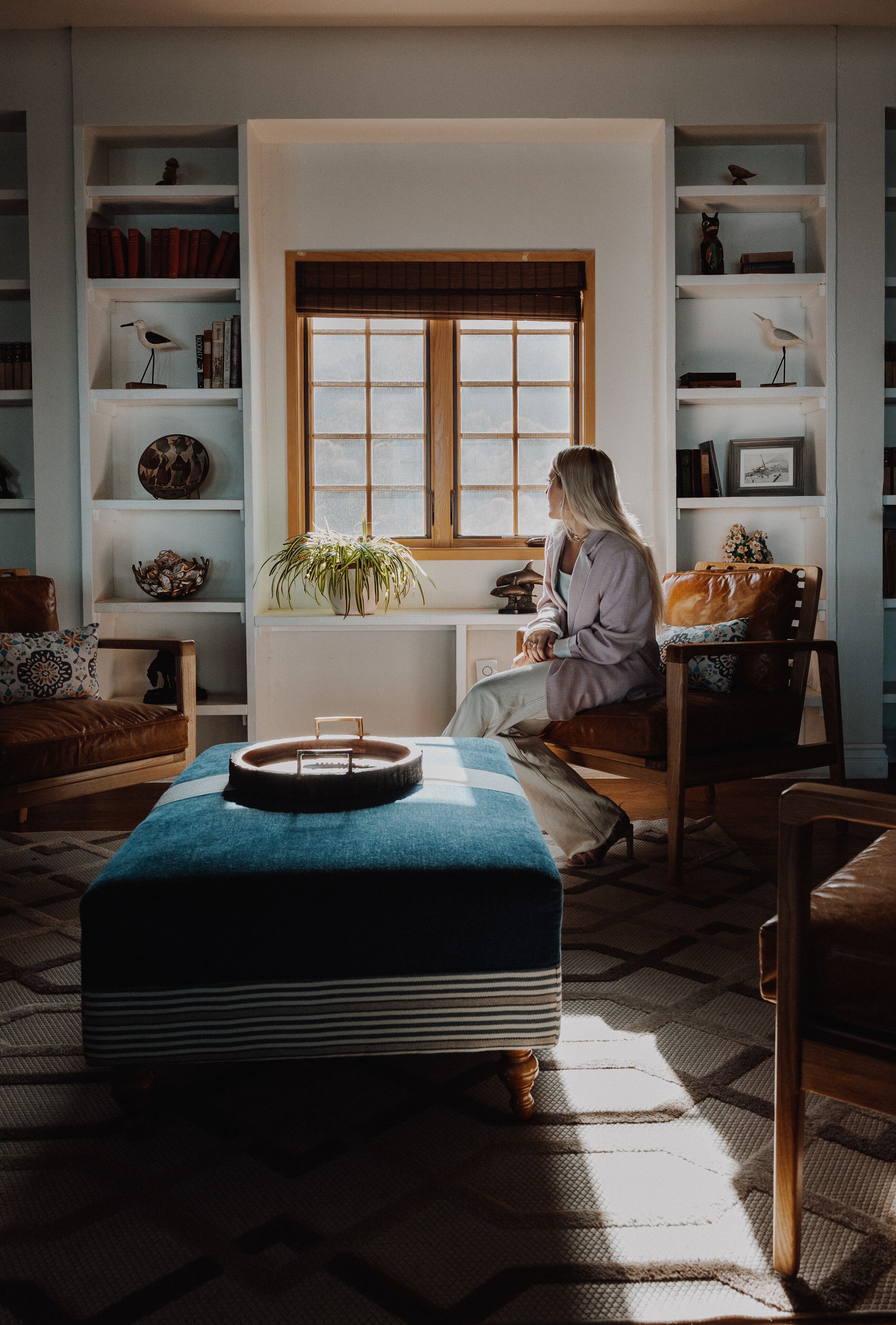 A cozy living room with a large window, a bookshelf, a brown leather sofa, and a blue ottoman.