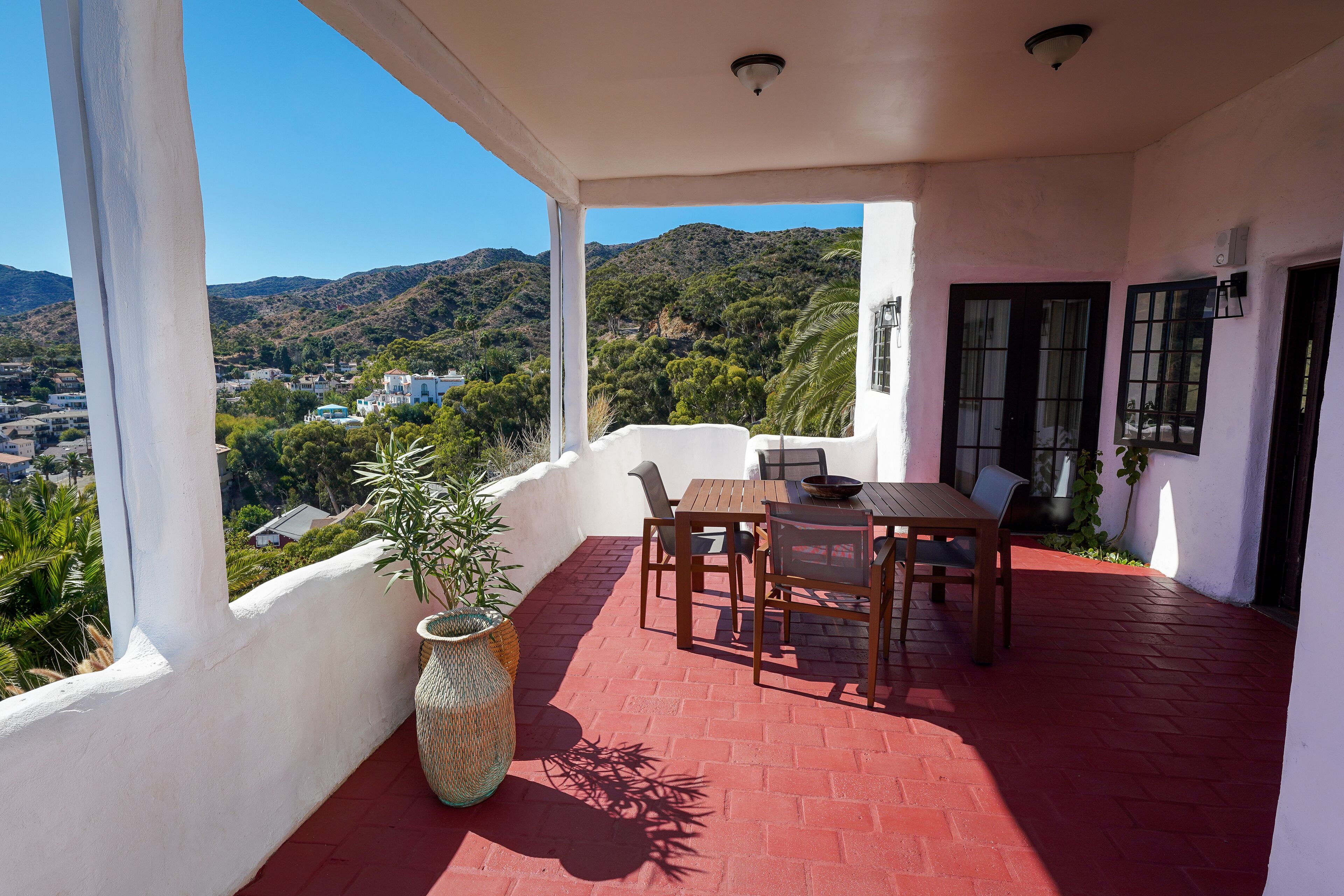 A covered patio with a table and chairs, a potted plant, and a view of a hillside with buildings.