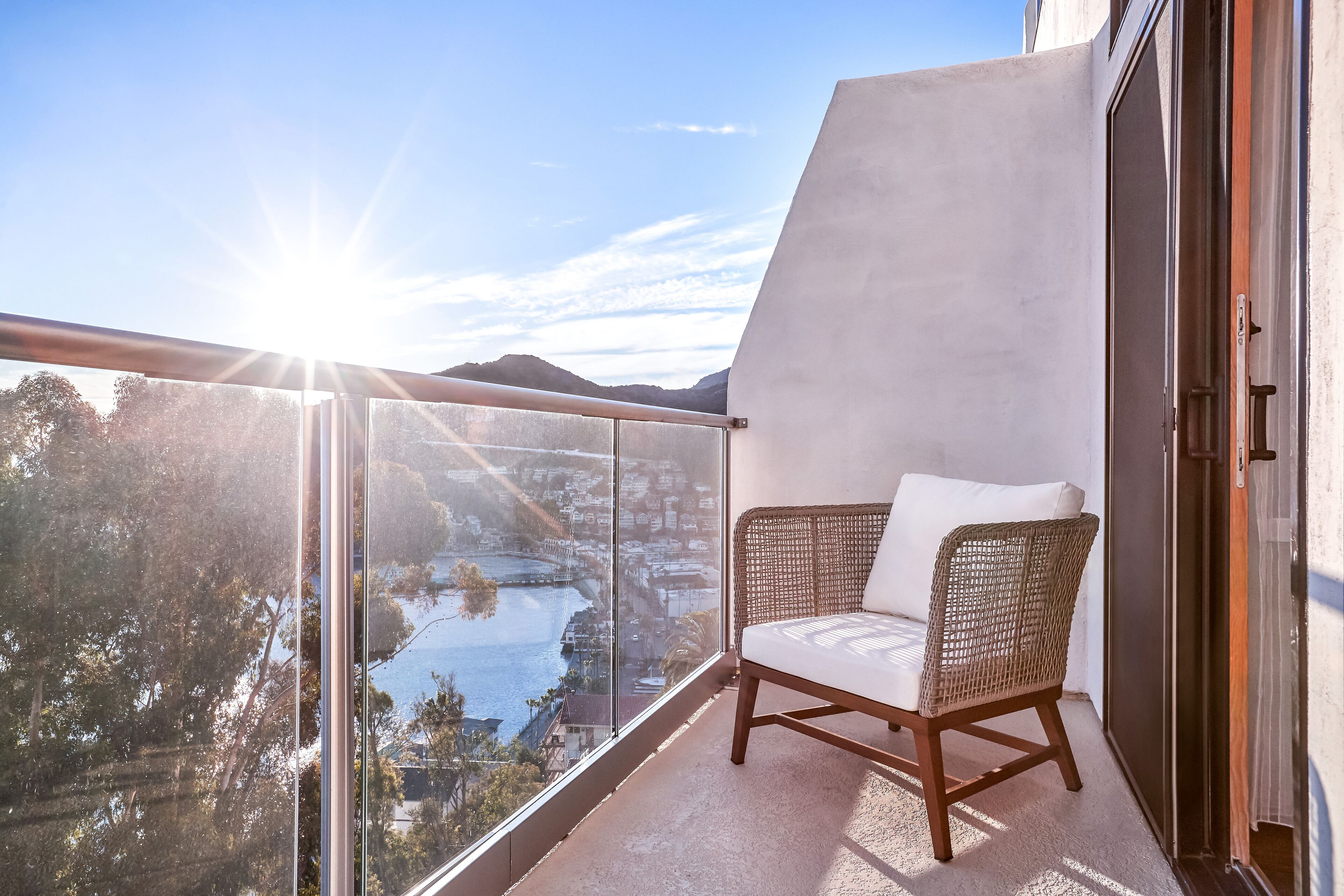 Ein Balkon mit einem Korbstuhl, einer Glastür und Blick auf eine Stadtlandschaft und Wasser.