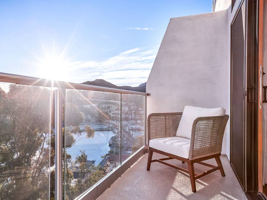 Ein Balkon mit einem Korbstuhl, einer Glastür und Blick auf eine Stadtlandschaft und Wasser.