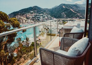 Un balcón con sillones de mimbre y baranda de cristal con vistas a un puerto deportivo con barcos y un paisaje montañoso.