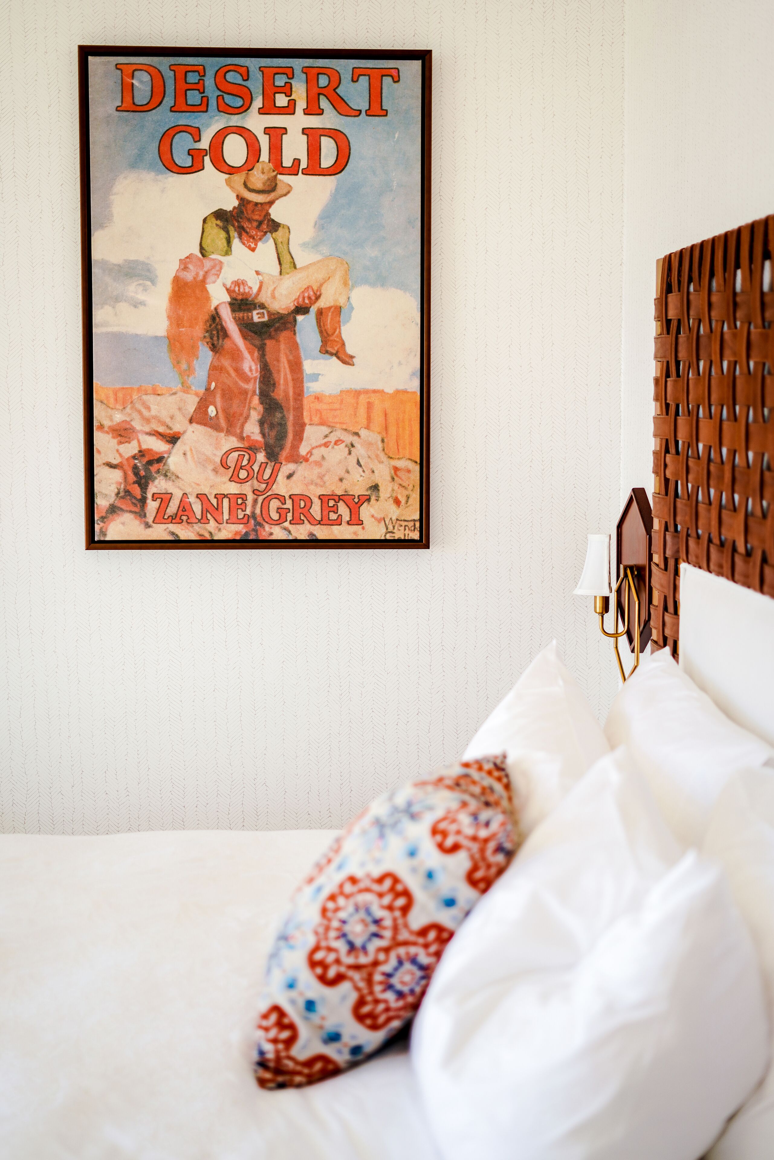 Et hotelværelse med et indrammet plakat af 'Desert Gold' af Zane Grey, en seng med hvide og mønstrede puder, og en træsenggavl.